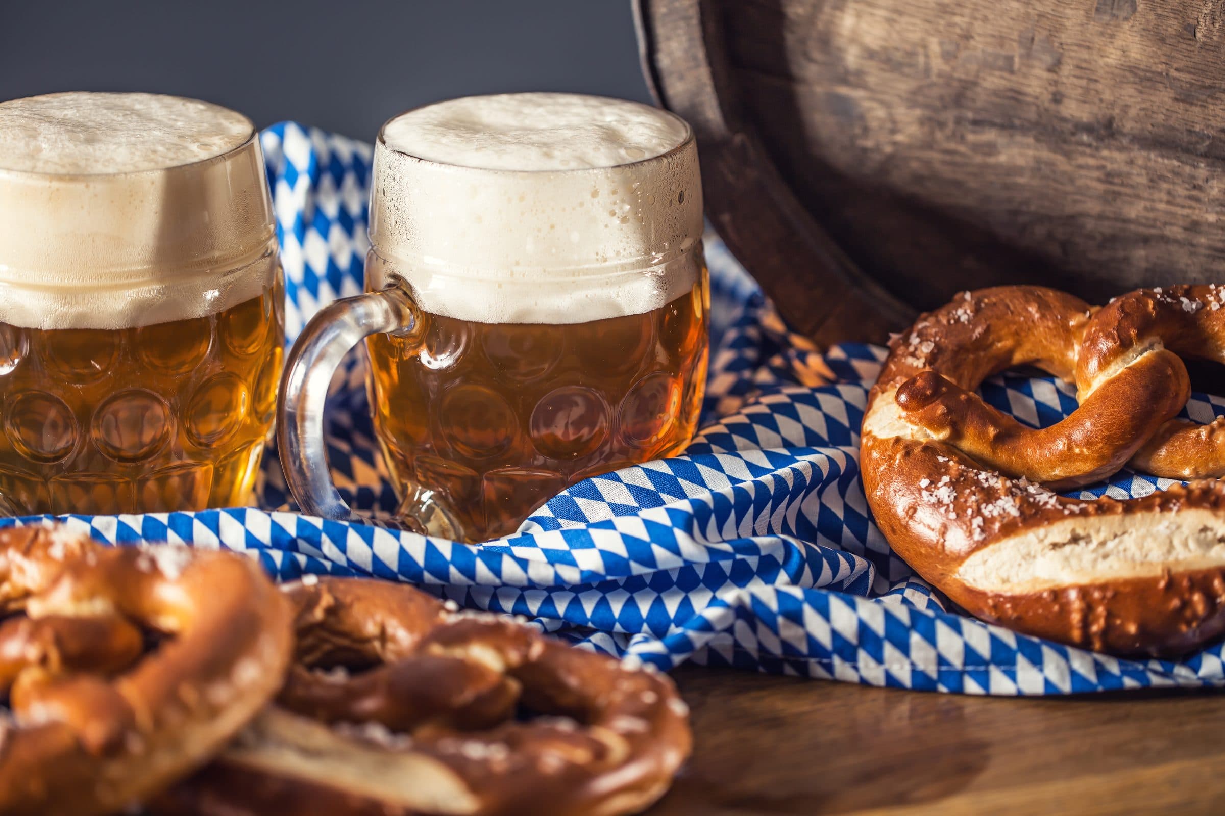 Zwei Maßkrüge Bier mit dicker Schaumkrone nebeneinander vor einem blau-weiß gerauteten Tuch, davor mehrere Laugenbrezeln mit grobem Salz auf einem Holztisch, rechts im Hintergrund ein altes Holzfass.